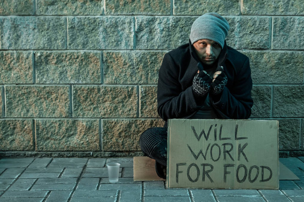 A man, homeless, a man asks for alms on the street with a sign will work for food. Concept of homeless person, addict, poverty, despair. - Photo, image