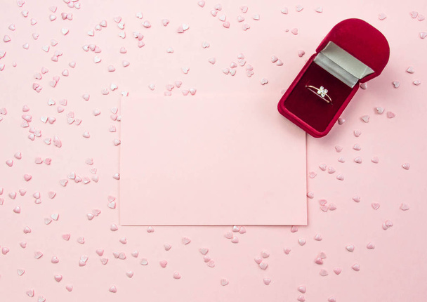 Wedding ring in a box and a pink sheet for writing on a pink background and decorative hearts. Marriage proposal concept, Valentines Day, International Womens Day. Flat lay, top view - Φωτογραφία, εικόνα