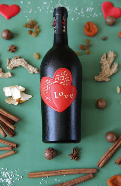  Flat lay. Still life with a bottle of wine with a red heart on a bottle with an inscription about love on a green background with spices and dried fruits for mulled wine Christmas Valentine's Day Love. Post card. Pattern. Vertical photo. - Photo, Image