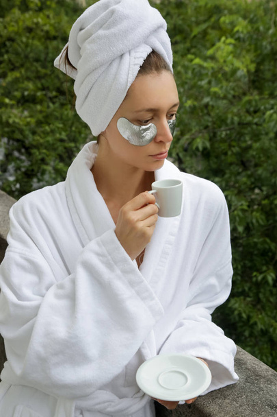 Γυναίκα με ένα μπουρνούζι και τυλιγμένο πετσέτα κεφαλής απολαμβάνοντας το πρωινό της στο μπαλκόνι, έχοντας ομορφιά κάτω από τα μάτια περιποίηση και πίνοντας καφέ - Φωτογραφία, εικόνα