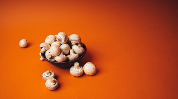 Pilze in einer Holzschale auf orangefarbenem Hintergrund. Der kleine weiße Champignon in einem Teller und in seiner Nähe verstreut. - Foto, Bild
