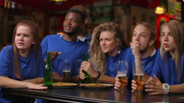 青いTシャツを着た男女のファンのグループは、バーで叫び声をあげてビールを飲んで喜びます。アフリカ系アメリカ人とヨーロッパ人 - 映像、動画