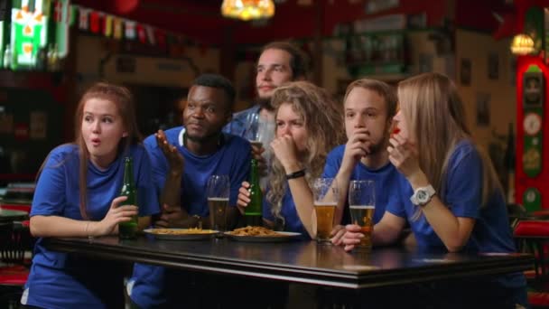 A multi-ethnic group of friends of fans in blue t-shirts will watch a match on TV in a bar, watch a tense exciting moment and celebrate a goal scored. Hugging, having fun, and making a glass of beer - Footage, Video
