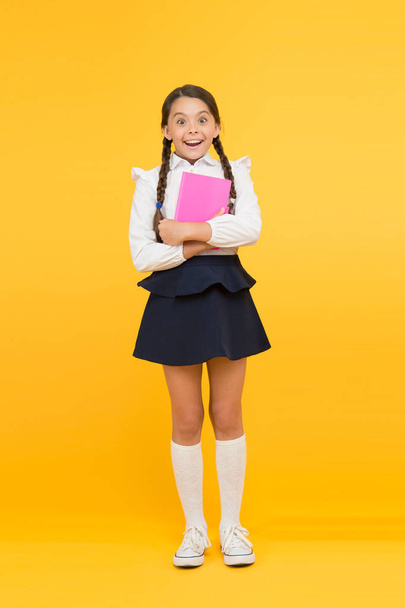 Exciting literature. Girl child hold book stand yellow background. Schoolgirl studying textbook. Kid girl school uniform happy face read book studying. Excited about knowledge. Enjoy studying - Photo, image