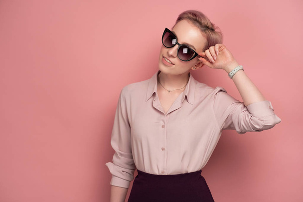 A girl with short pink hair in a blouse and skirt, smiling, looking at the camera in sunglasses, standing on a pink background. - Foto, Imagem