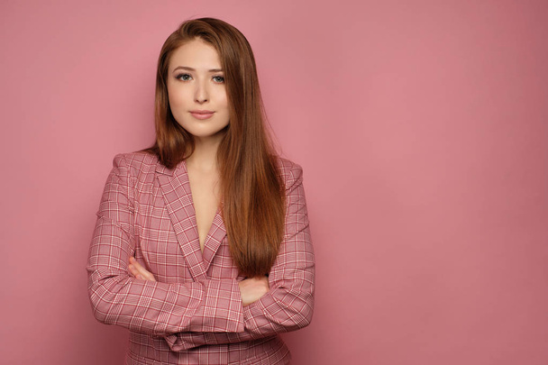 Red-haired girl in a business suit stands on a pink background with her arms folded and smiling looking at the camera. - Foto, Imagen