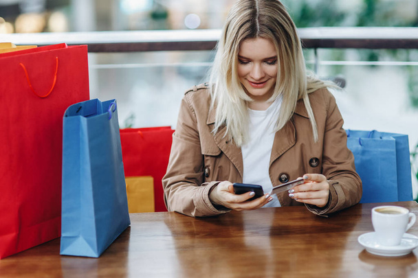 The girl spent a lot of money. Blonde woman in brown cloak with shopping bags in cafe at shopping mall. Lady buying online with a credit card and smartphone. Coffee cup on table - Photo, image