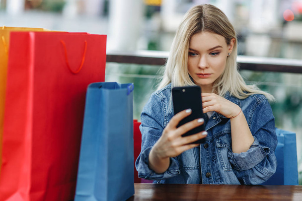 Woman in a jeans jacket with shopping bags sitting in cafe at shopping mall. Girl playing with mobile phone in her hand. Buying too much concept. Concept - Relaxing after shopping - Zdjęcie, obraz