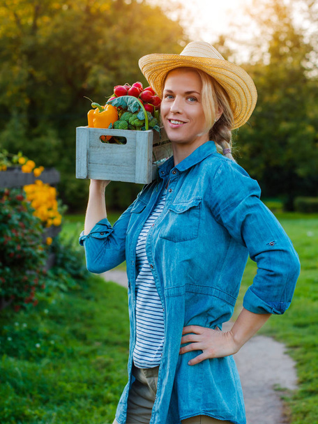 Jonge 30-35 jaar oud mooie vrouw boer met hoed met doos verse ecologische groenten op tuin achtergrond bij zonsondergang. - Foto, afbeelding