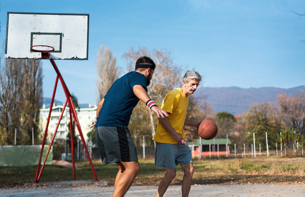 Senior man playing basketball with his son in a park - Фото, изображение