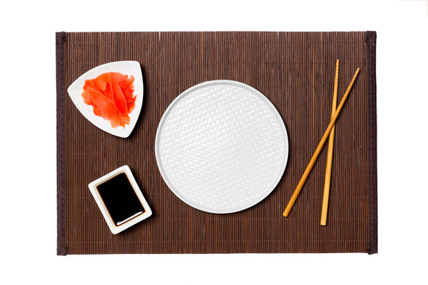 Lege ronde witte plaat met eetstokjes voor sushi, gember en sojasaus op donkere bamboe mat achtergrond. Bovenaanzicht met Kopieer ruimte voor uw ontwerp - Foto, afbeelding