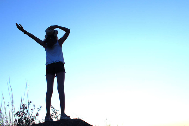 Silhueta de uma jovem adolescente usando um chapéu para trás, olhando para o pôr do sol e de mãos erguidas. Pessoas ao ar livre, viagens, conceito de paisagem
.  - Foto, Imagem