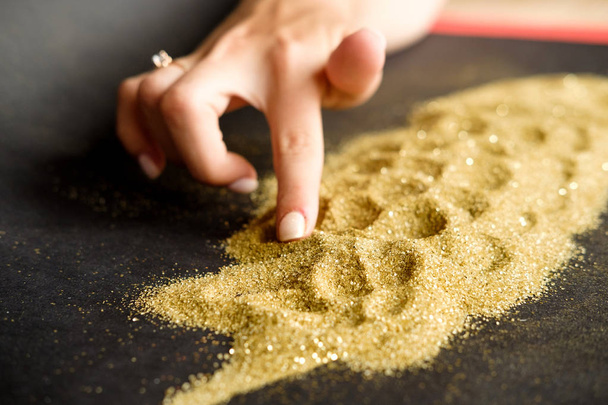 Kobieta rysuje na złotym błyszczącym piasku na czarnym tle. Wydobycie złota - Zdjęcie, obraz