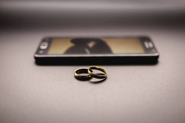 Concept van liefhebbers of huwelijkse verraad, ontrouw. Paar ringen naast een smartphone, met een afbeelding van een wazig paar zoenen op het scherm. - Foto, afbeelding