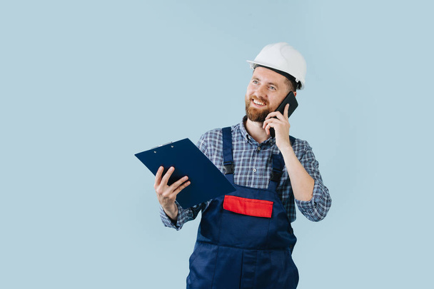 Happy construction worker in a white helmet and blue overalls with a phone - Foto, Bild