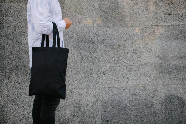 Young man holding black textile eco bag against urban city background. . Ecology or environment protection concept. Black eco bag for your design or logo mock up - Foto, imagen