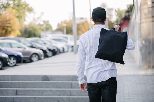 junger Mann mit schwarzer Textil-Öko-Tasche vor städtischem Hintergrund. . Ökologie oder Umweltschutzkonzept. schwarze Öko-Tasche für Ihr Design oder Logo-Attrappe - Foto, Bild