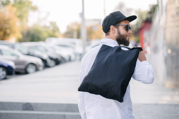 Νέος άνθρωπος που κατέχει μαύρη υφαντουργική τσάντα Eco κατά του αστικού ιστορικού της πόλης. . Οικολογία ή έννοια προστασίας του περιβάλλοντος. Μαύρη τσάντα Eco για το σχεδιασμό ή το λογότυπο σας - Φωτογραφία, εικόνα