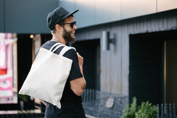 Young man holding white textile eco bag against urban city background. . Ecology or environment protection concept. White eco bag for mock up. - 写真・画像