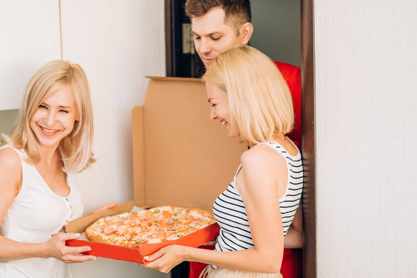 Twee jonge Kaukasische vrouw ontvangen pizza van delivery man thuis - Foto, afbeelding