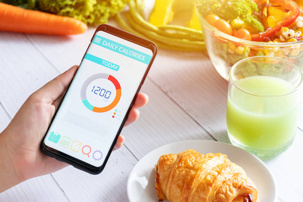 Calories counting and food control concept. woman using Calorie counter application on her smartphone with salad , vegetable, juice and croissant on dining table - Fotoğraf, Görsel