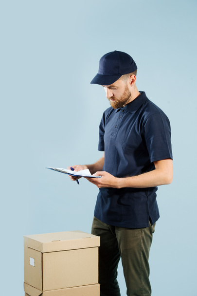 Delivery man is checking his orders list while standing next to stack of boxes - Fotó, kép