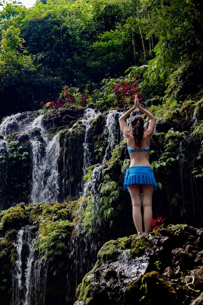 若い白人女性が瞑想し、滝でヨガを練習しています。後ろから見る。ナマステ・ムドラバニュワナアメルタ滝ワナギリ、バリ、インドネシア.  - 写真・画像