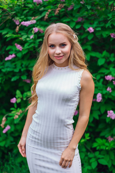 Portrait of a charming blond woman wearing beautiful white dress standing next to lilac bush. - 写真・画像