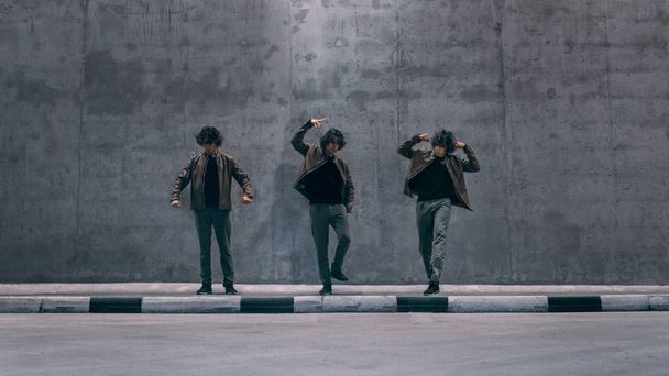 Tiro de exposición múltiple de un joven hipster clonado fresco con soportes de pelo largo en la danza posa en una calle junto a una gran pared de hormigón. Lleva una chaqueta de cuero marrón.
. - Foto, imagen