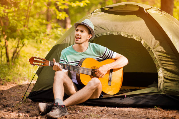 幸せな若い男のキャンプや森の夕日を背景にリラックスするためのギター楽器の音楽を促進します。ワイルダネストラベルライフスタイルコンセプト. - 写真・画像