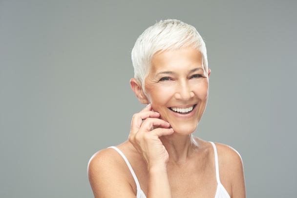 Beautiful smiling senior woman with short gray hair posing in front of gray background. Beauty photography. - Photo, Image