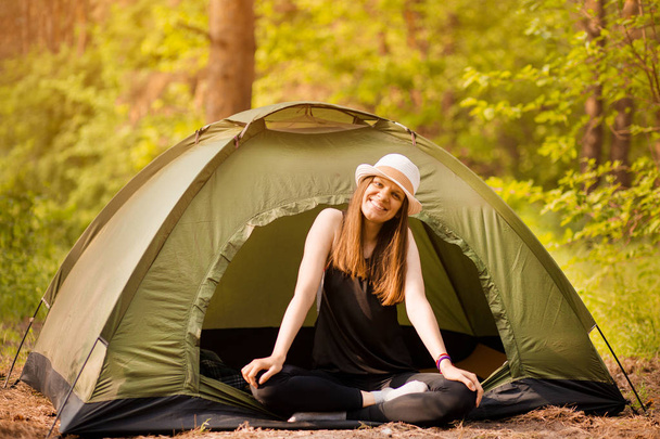Gelukkige vrouw in de hoed zit in een tent rond bos onder zonsondergang licht hemel genieten van vrije tijd en vrijheid. - Foto, afbeelding