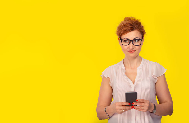 Daydreaming, thoughtful with a smartphone. Happy woman looking to side thinking, holding a phone. Isolated yellow studio background with copy space. - 写真・画像