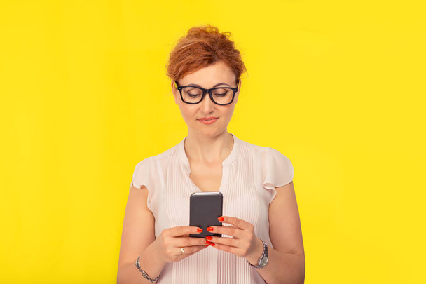 Closeup portrait of a beautiful woman holding using looking at smart phone, girl wearing eyeglasses and nude color casual shirt, with retro curly hair style up, cutout isolated on a yellow background - 写真・画像