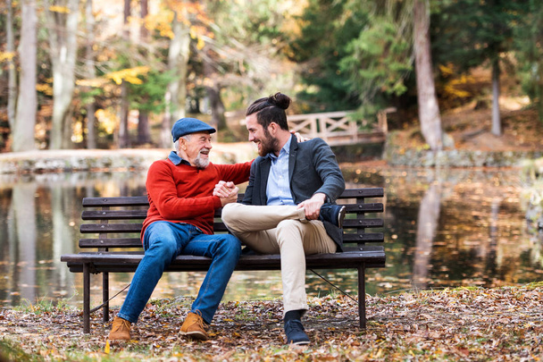Senior father and his son sitting on bench by lake in nature, talking. - Photo, Image