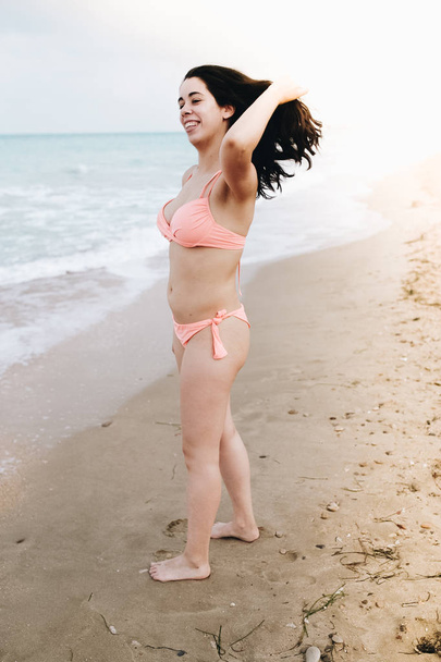 Happy young woman with in pink bikini touching her hair in blue clear water on sand beach in a cloudy day. Travel concept. - 写真・画像