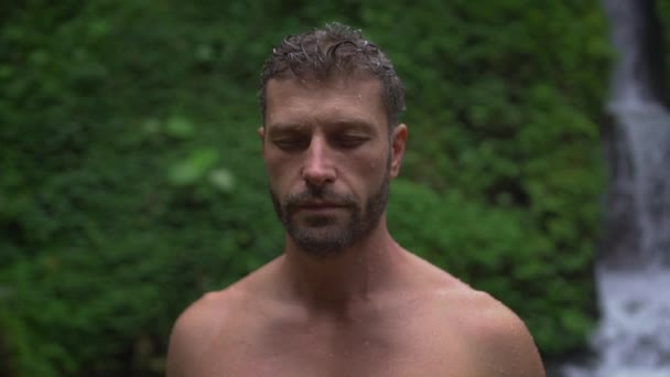 Handsome man with wet face and hair stands near the waterfall with closed eyes - Imágenes, Vídeo