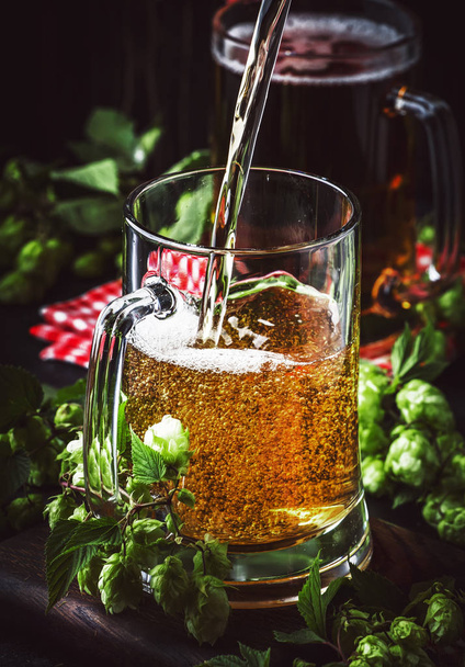 Large mugs with german light beer and fresh hops, pours into glass, dark table, selective focus - Foto, Imagem
