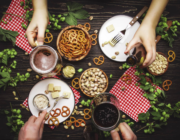 Beer summer party: dark and light beer, appetizers, salty snacks, hop cones, plates and glasses in the hands of people. Top view, dining wooden table background, people relaxing on day off concept - 写真・画像