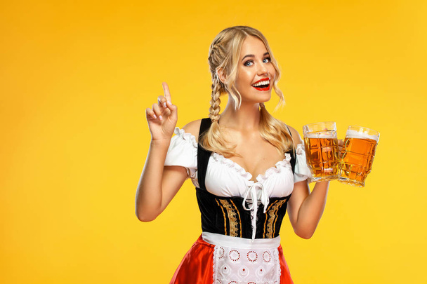 Young sexy Oktoberfest girl waitress, wearing a traditional Bavarian or german dirndl, serving big beer mugs with drink isolated on yellow background. Woman pointing to looking up. - Fotografie, Obrázek