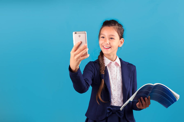 Cute cheerful smiling school girl in formal dressing with exercise book in her hand looking to screen of smartphone, shoot selfie, blue background. Online education, communication with tutor concept - Фото, изображение