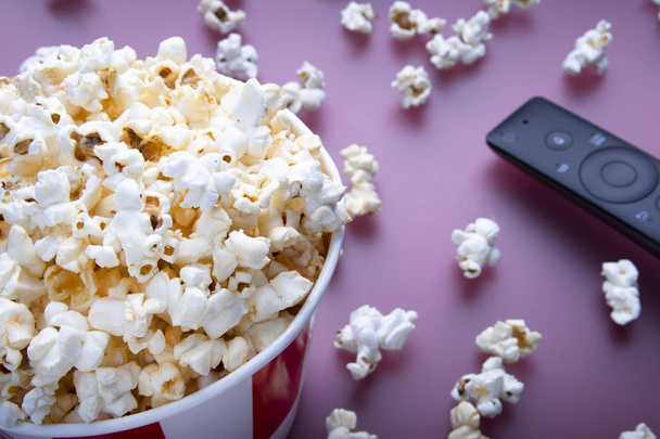 Close-up of remote control and striped bowl of popcorn on red background. The concept of watching a movie, TV, DVD. Entertainments - Photo, Image