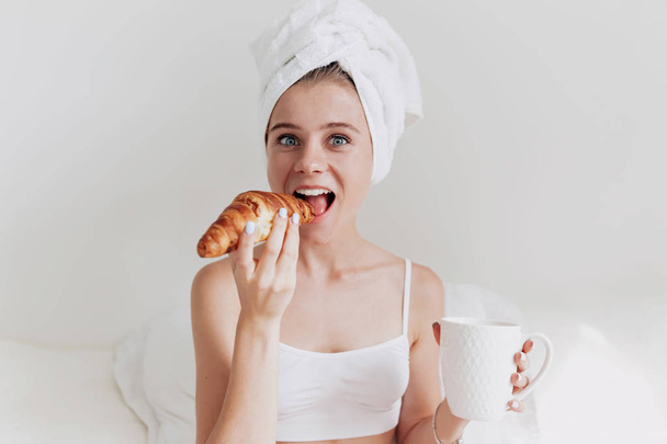 Izgatott vicces hölgy törülközővel a fejét csípős reggel croissant és kávét iszik, fehér ágyban - Fotó, kép