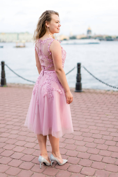 Young curly Caucasian woman in rose pink dress turned her back on embankment. - Foto, imagen