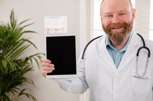 lächelnder erwachsener Arzt, der ein digitales Bild oder einen Bericht auf einem Tablet zeigt - Foto, Bild