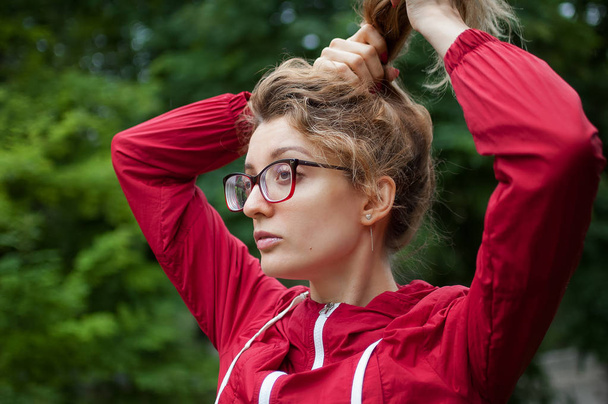 Portrait of blonde girl with hipster eyeglasses in dark red jacket making ponytail outdoors in the urban park - Photo, Image