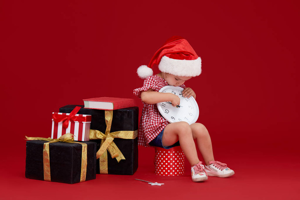 Happy child girl in santa hat near the gift boxes. Kid reading a book at Christmas night. Happy xmas and New Year, winter holiday concept with copy space for text on red background - Photo, image