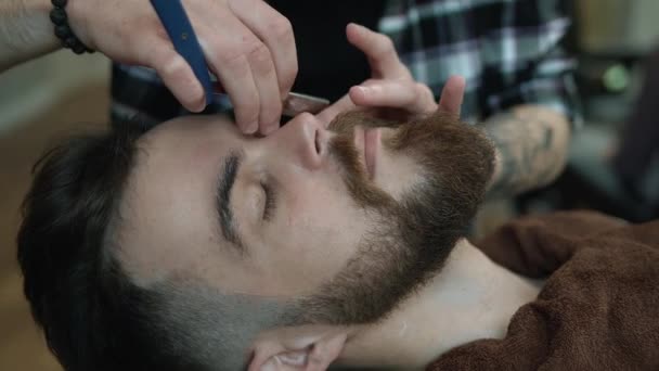 Barber shaves men with a long beard with straight razor blade in s hair salon or barbershop. Mans haircut and shaving at the hairdresser, barber shop and shaving salon. - Filmati, video