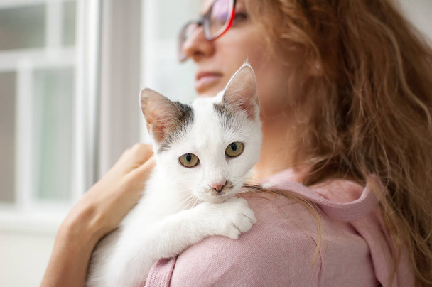 Attractive girl in pink clothes and eyeglasses with withe cat in her arms standing near the window at home. Pet owner concept - Fotó, kép