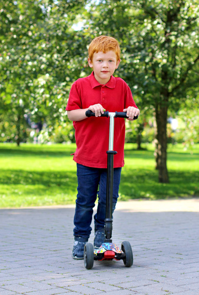 Child learn to ride scooter in a city park on sunny summer day. Cheerful little boy with ginger hair posing with a scooter. Active leisure and outdoor sport for child - Foto, Imagem